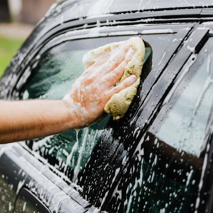 Close-up of a person washing a car window with a sponge and soap suds outside.